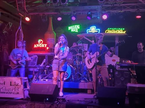 A lively band performs on stage with colorful lights and neon signs in the background, creating a vibrant atmosphere.