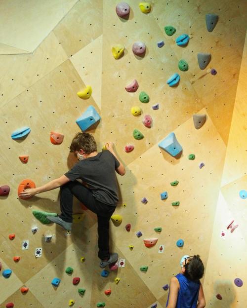 Two children climbing a colorful indoor bouldering wall, one scaling the wall while the other watches.