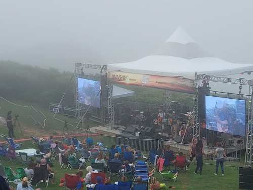 A foggy outdoor concert scene with a stage, large screens, and an audience seated on lawn chairs.
