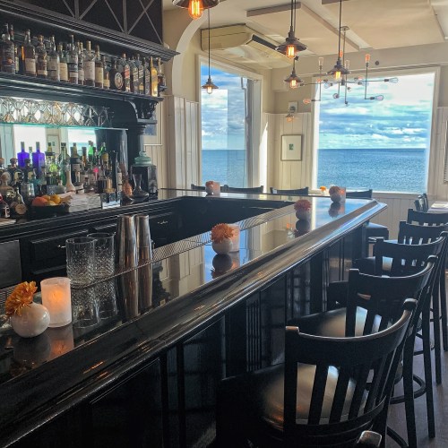 A stylish bar with a black countertop, flowers, and a view of the ocean through large windows.