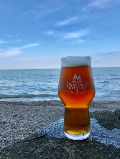 A glass of amber beer sits on a rock by the beach, with calm waters and a blue sky in the background.