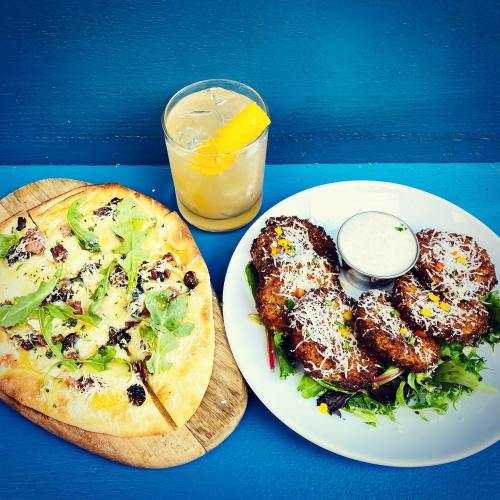 A wooden board with a pizza topped with greens, and a plate of fritters with a dipping sauce, alongside a cocktail.