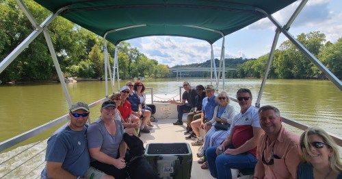 A group of people sitting on a boat, enjoying a scenic river view surrounded by trees and a bridge in the background.