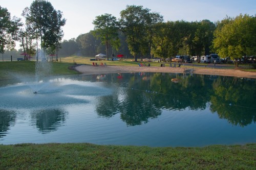 A serene pond surrounded by trees, with a fountain and camping area in the background on a clear morning.