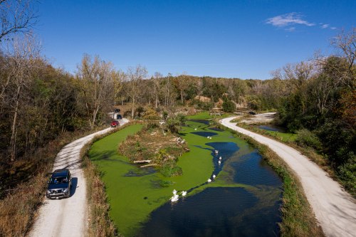 Aerial view of two winding roads beside a green, algae-covered river, surrounded by trees under a clear blue sky.