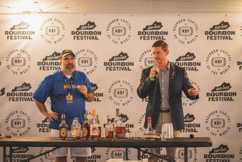 Two men stand behind a table with bourbon bottles, speaking at the Kentucky Bourbon Festival under a branded backdrop.