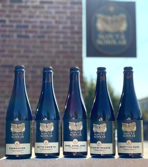 Five bottles of Scout & Scholar beer lined up in front of a brick wall, showcasing their unique labels.