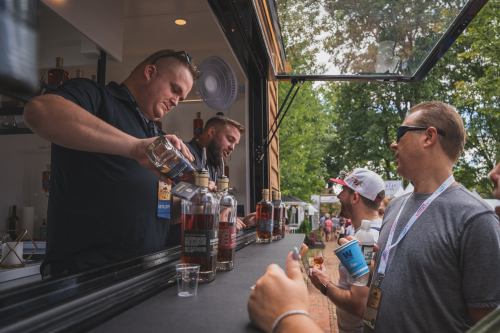 Two bartenders serve drinks at a festival booth while attendees enjoy their beverages in a lively outdoor setting.