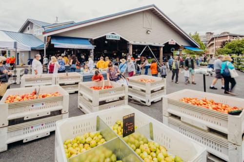 A bustling market scene with people shopping, featuring crates of red tomatoes and green apples outside a building.