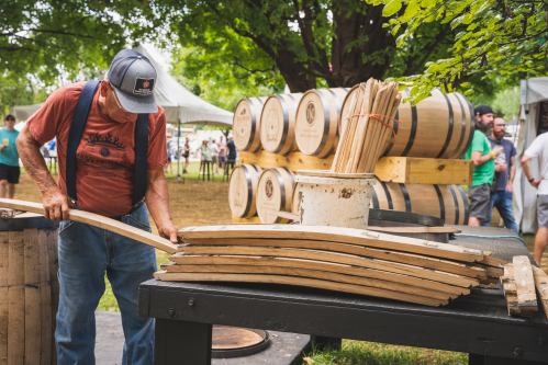 A man in a cap works with wooden staves at a festival, with barrels and people in the background.