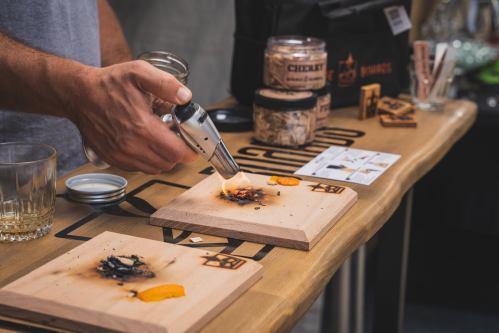 A person uses a torch to char wood on a wooden board, with ingredients and tools visible on a table.