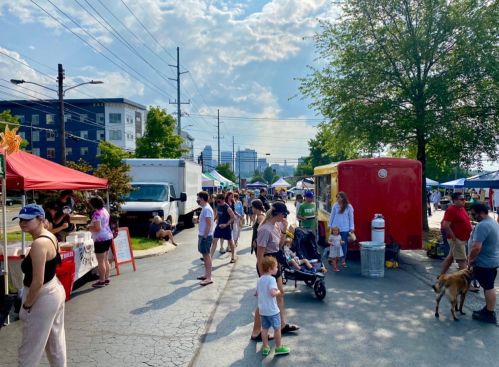 A bustling outdoor market scene with people browsing stalls, food trucks, and children playing under a sunny sky.
