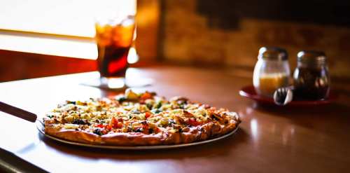 A freshly baked pizza topped with vegetables on a wooden table, with a drink and condiments in the background.