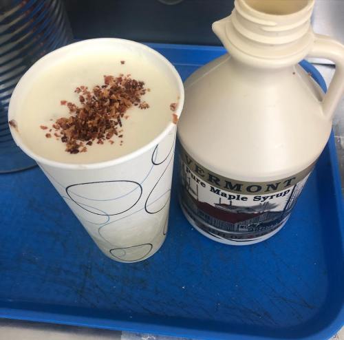 A cup of creamy beverage topped with bacon bits, next to a bottle of Vermont maple syrup on a blue tray.