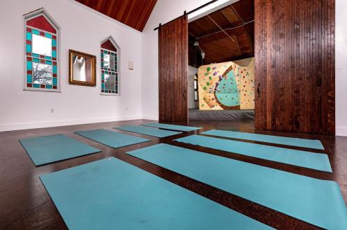A spacious room with turquoise yoga mats arranged on the floor, stained glass windows, and a climbing wall in the background.