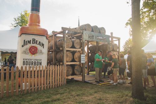A large Jim Beam bottle stands beside a display of bourbon barrels and a crowd at an outdoor event.