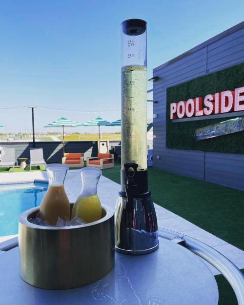A tall drink dispenser filled with a green beverage next to two glass pitchers of juice by a poolside area.