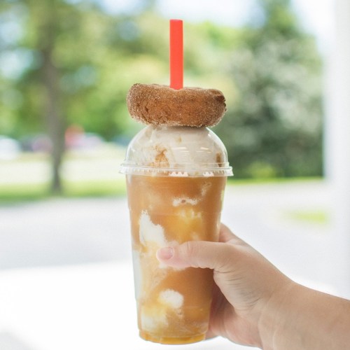 A hand holding a clear cup of iced drink topped with ice cream and a donut, with a red straw.