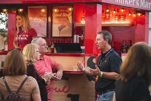 A man talks animatedly to a couple at a Maker's Mark booth, with people enjoying drinks in a lively outdoor setting.
