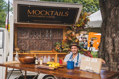 A vendor at a market stand offers mocktails and cold-brew coffee, surrounded by autumn decorations and fresh lemons.