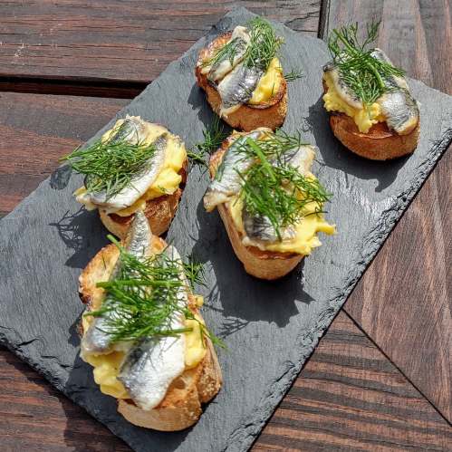 Five toasted bread slices topped with scrambled eggs, anchovies, and fresh dill, served on a slate platter.