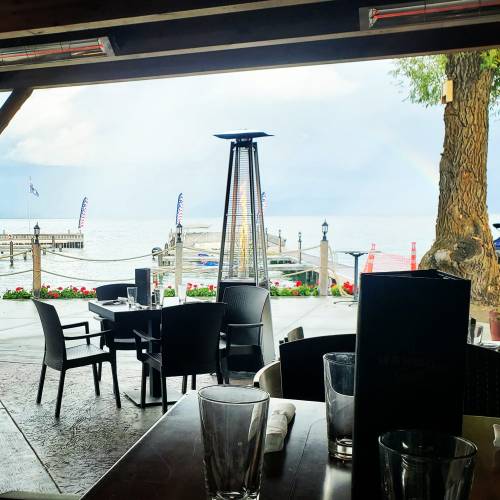 View from a restaurant with empty tables, a lake, and a rainbow in the distance under a cloudy sky.