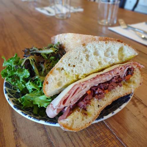A sandwich with layers of meat and cheese, served with a side of fresh mixed greens on a wooden table.