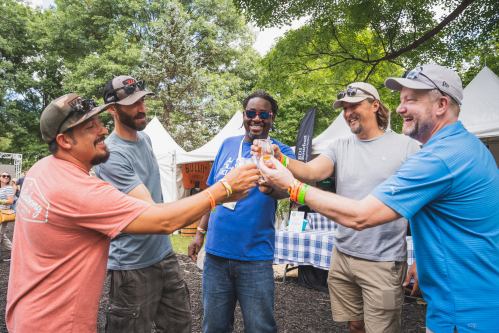 Five men smiling and toasting with drinks at an outdoor festival, surrounded by tents and greenery.