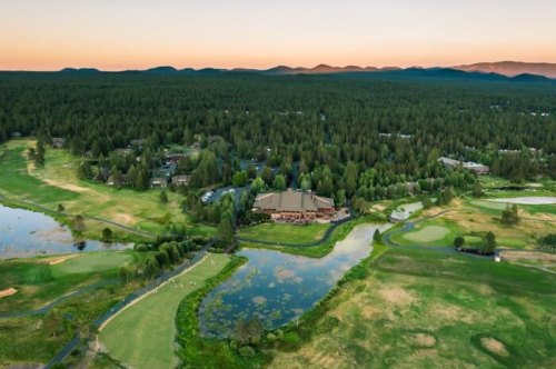 Aerial view of a golf course surrounded by lush greenery and water features, with mountains in the background at sunset.