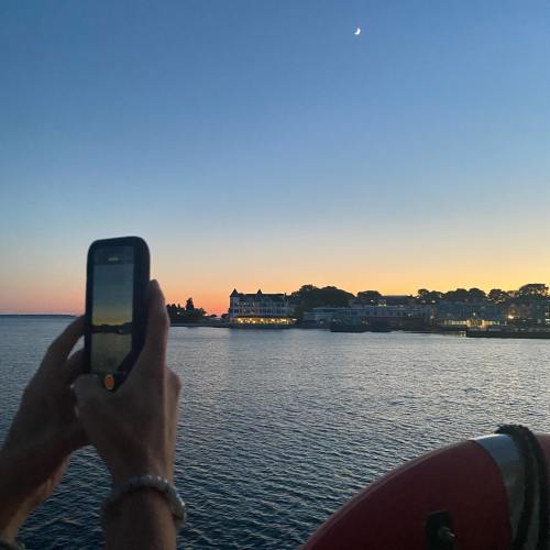 A person holds a phone, capturing a sunset view over water with a distant shoreline and a crescent moon in the sky.