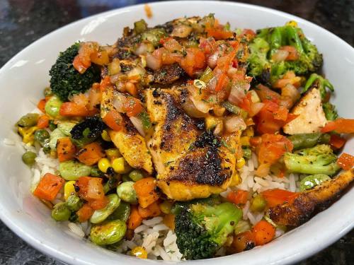 A bowl of rice topped with grilled chicken, fresh salsa, and a mix of colorful vegetables like broccoli, corn, and peppers.