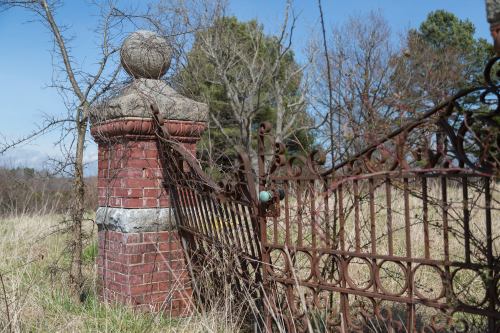 An old, rusted gate with a brick pillar, surrounded by overgrown grass and trees under a clear blue sky.