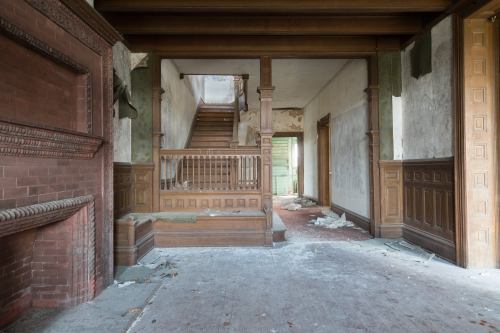 Abandoned interior of a house with a staircase, wooden paneling, and debris scattered on the floor.