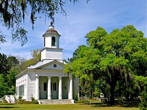 A historic white church with a bell tower, surrounded by lush green trees and Spanish moss under a clear blue sky.