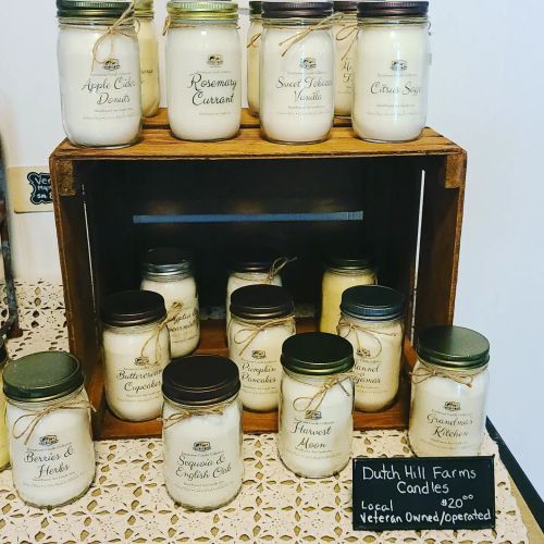 A display of various scented candles in jars, labeled with names like "Apple Crisp" and "Rosemary," on a wooden shelf.