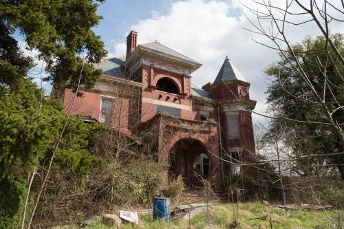 Abandoned red brick mansion surrounded by overgrown vegetation and trees under a cloudy sky.
