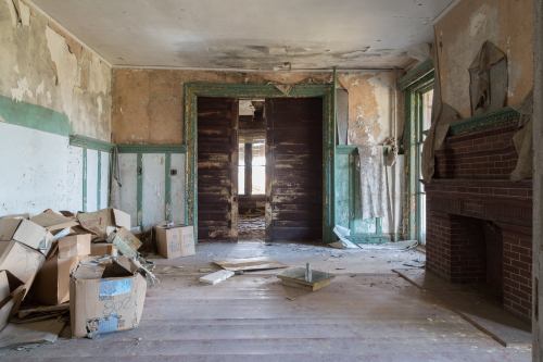 An abandoned room with peeling walls, scattered boxes, and a fireplace, showcasing signs of neglect and decay.