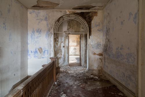 A dilapidated hallway with peeling blue wallpaper, debris on the floor, and a partially collapsed ceiling.