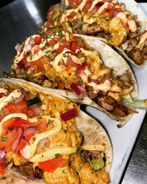 Three delicious tacos filled with grilled meat, lettuce, tomatoes, and drizzled with creamy sauce on a white plate.