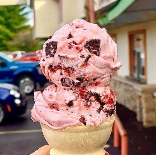 A close-up of a pink ice cream cone topped with chocolate chunks and cherry pieces, held against a blurred background.