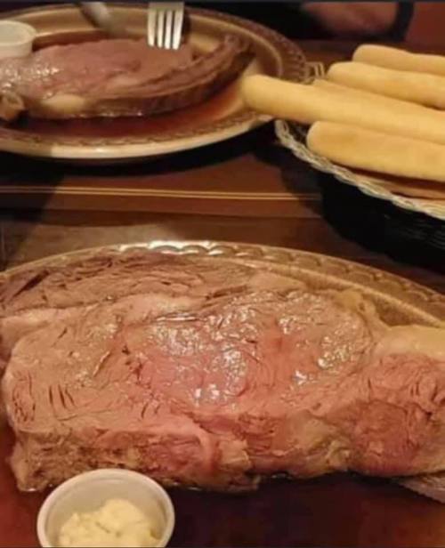 A large, juicy prime rib served with a side of creamy sauce and a basket of breadsticks in the background.