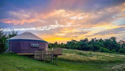 A yurt on a grassy hill at sunset, with colorful clouds and trees in the background.
