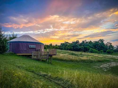 A yurt on a grassy field at sunset, with colorful clouds and trees in the background.