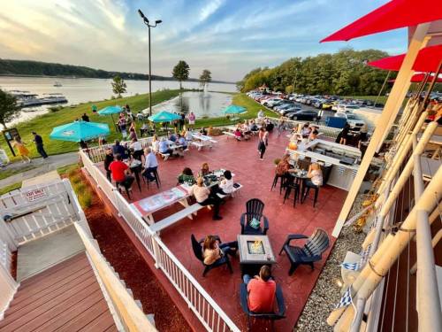 Outdoor dining area by a river, with people enjoying food and drinks, surrounded by greenery and parked cars.