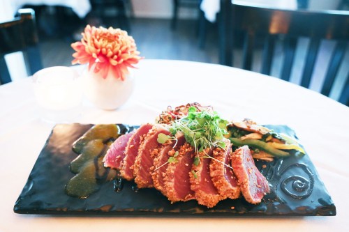 A beautifully plated dish of seared tuna with garnishes, served on a black slate, alongside a small flower vase.