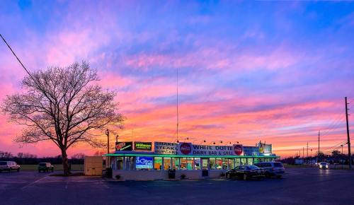 A colorful sunset over a diner with neon signs, surrounded by trees and parked cars.