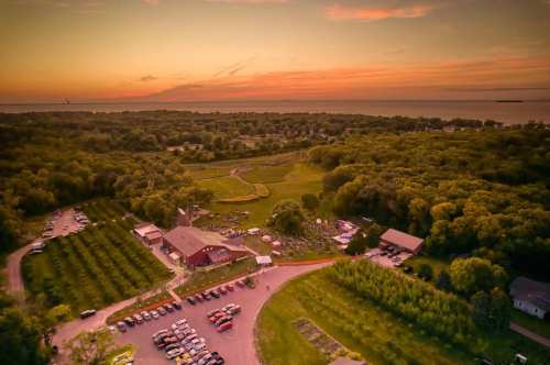 Aerial view of a scenic landscape at sunset, featuring a red barn, parking area, and lush greenery by the water.