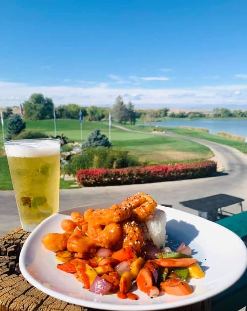 A plate of shrimp and vegetables with a beer, overlooking a scenic golf course and lake on a sunny day.