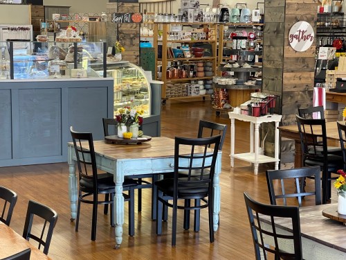 A cozy café interior featuring a wooden table with chairs, display cases, and shelves filled with kitchenware and decor.