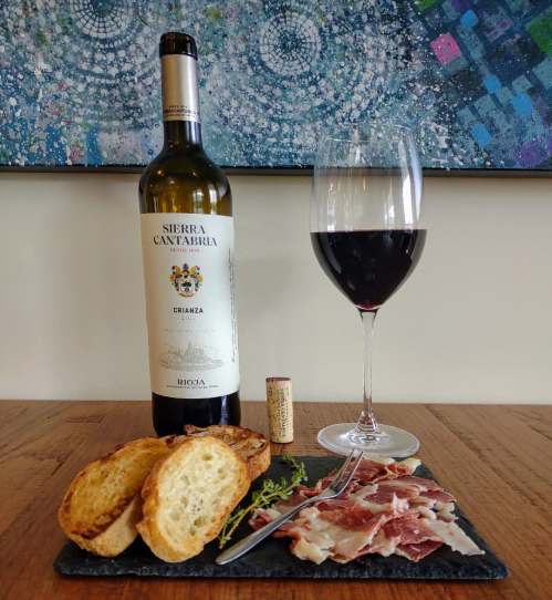 A bottle of red wine, a glass, sliced cured meat, toasted bread, and a cork on a slate board against a colorful background.